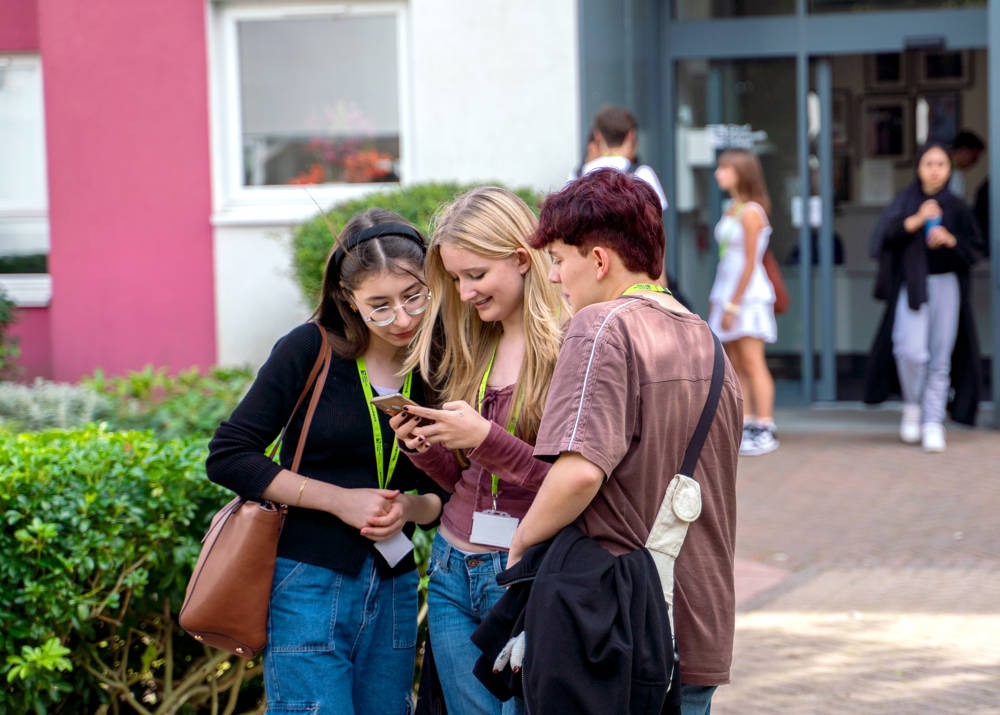 Three students looking at a friends phone outside the entrance of the Sixth Form Campus