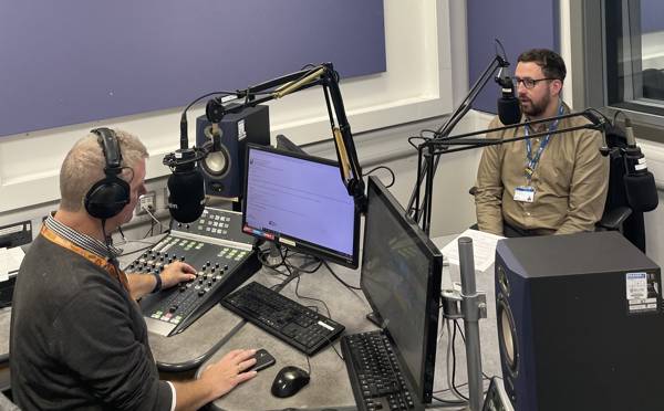 COPC Assistant Principal Lee McCarthy sits facing the camera at a DJ booth at Express FM, interviewed by Ian James.
